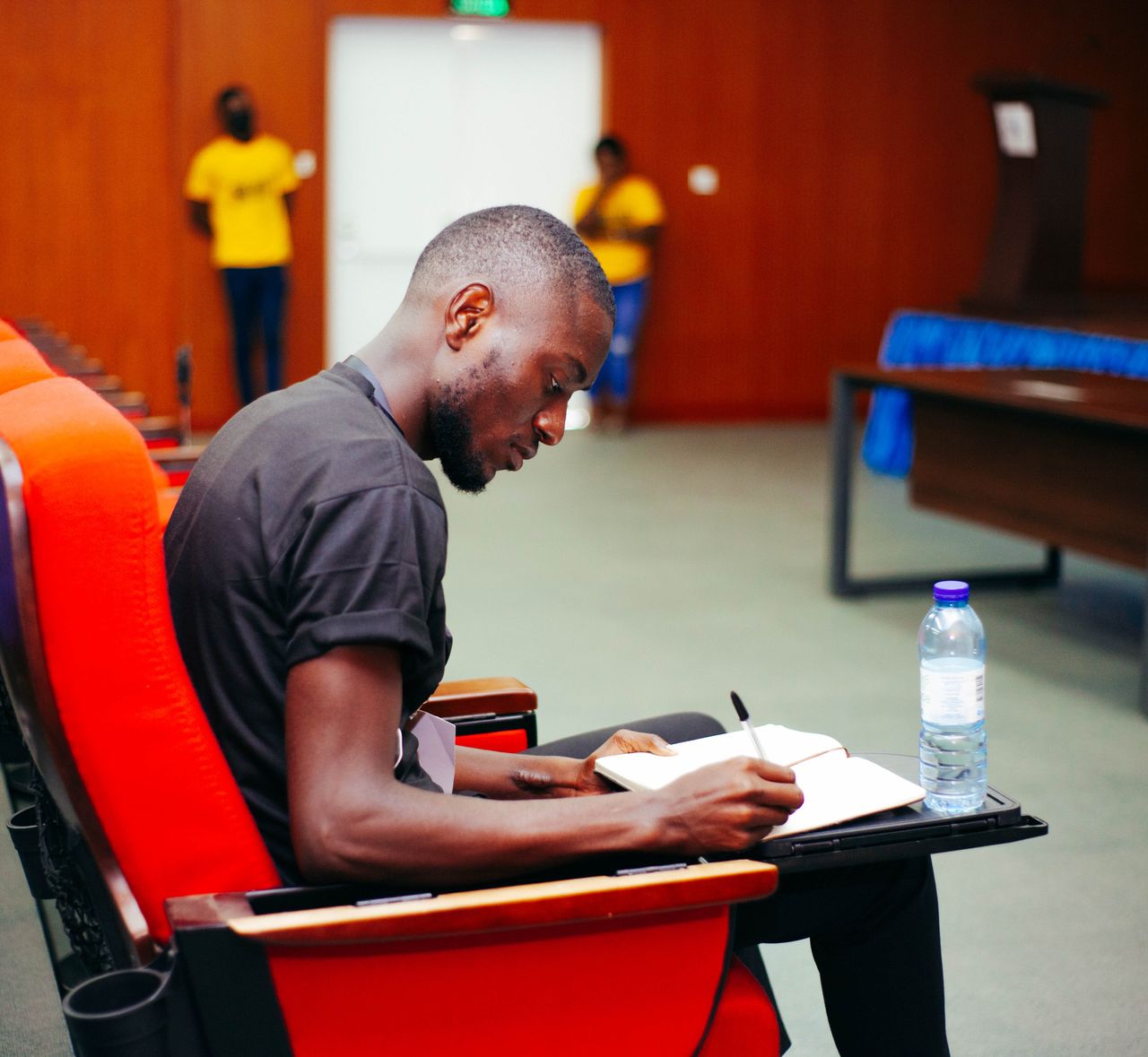 Black man sitting in a conference room, writing notes with a water bottle visible.