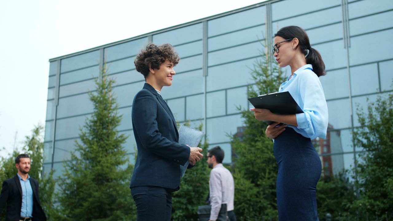 why-choose-us Two businesswomen engage in conversation outside a modern office building.